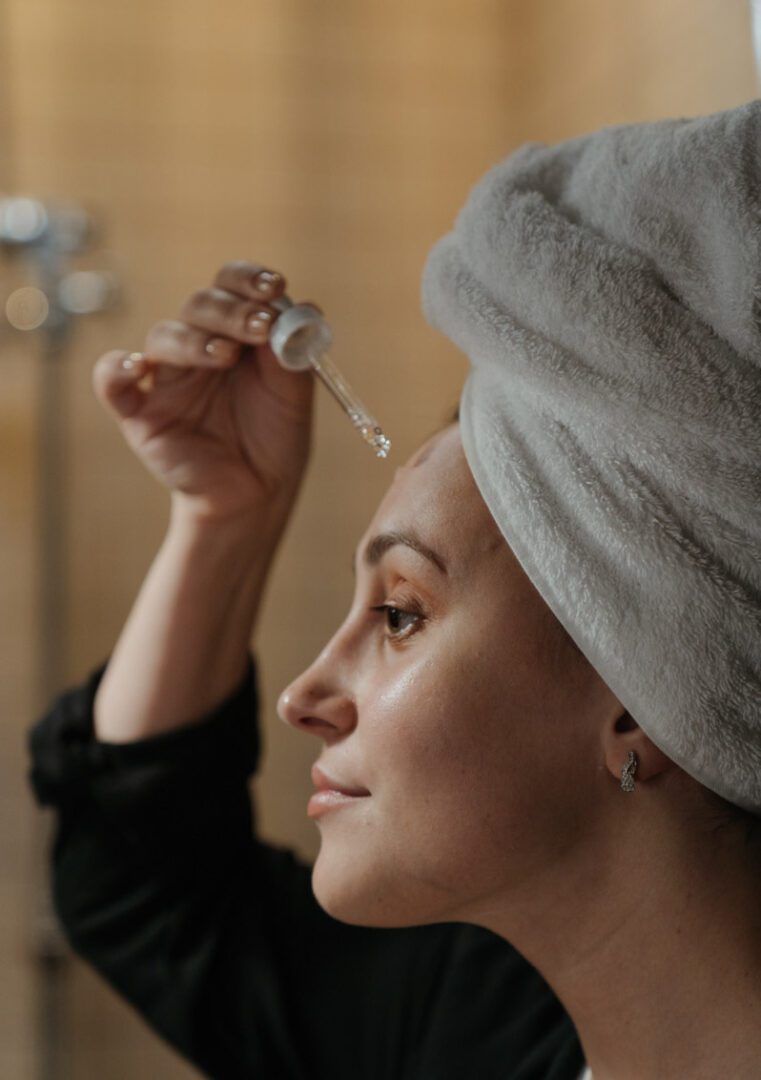 A woman with a towel wrapped around her head is putting lotion on her face.